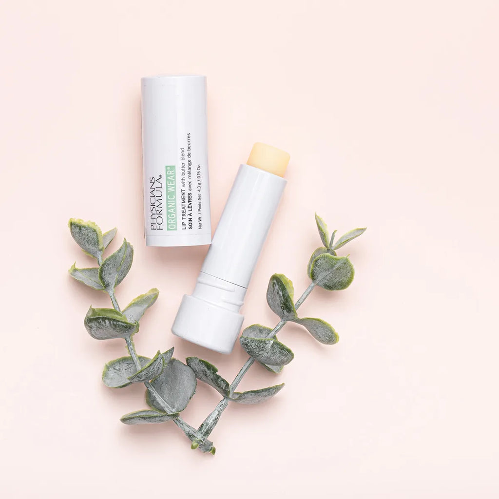 Physicians Formula Organic Wear Lip Treatment stick in white packaging, placed with green botanical accents on a soft pink background